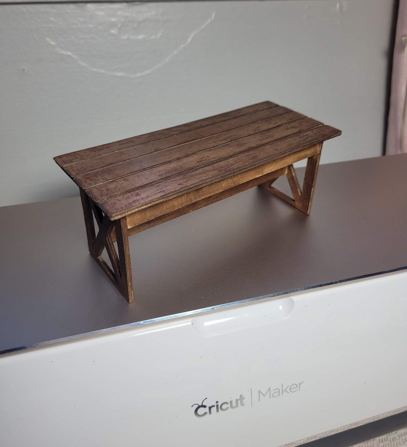 May include: A miniature wooden table with a brown stain finish. The table has a rectangular top with a simple design and a base with an X-shaped support.