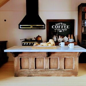 May include: Miniature kitchen scene with a marble countertop island and a wooden base. A black range hood hangs above a stove and a cutting board with bread. A chalkboard sign reads "Brewed Coffee served daily". Two white mugs sit on the counter.