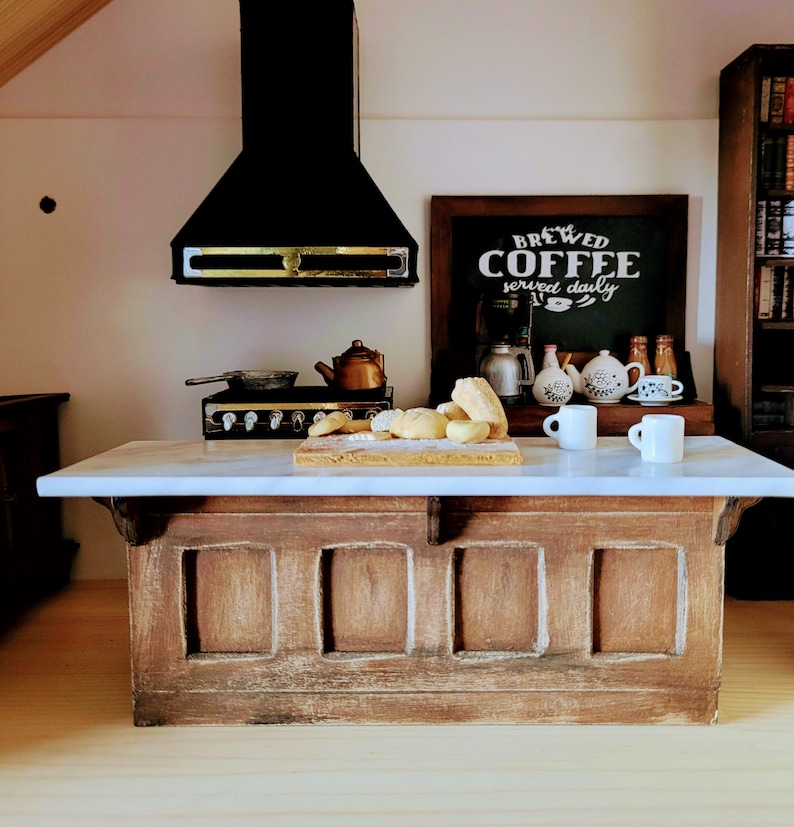 May include: Miniature kitchen scene with a marble countertop island and a wooden base. A black range hood hangs above a stove and a cutting board with bread. A chalkboard sign reads "Brewed Coffee served daily". Two white mugs sit on the counter.