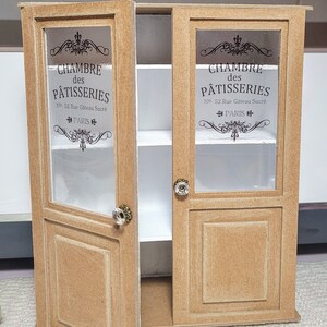 May include: A miniature wooden cabinet with two glass doors. The doors are open and feature a printed design that says "CHAMBRE des P&Acirc;TISSERIES No. 52 Rue G&acirc;teau Sucr&eacute; PARIS".