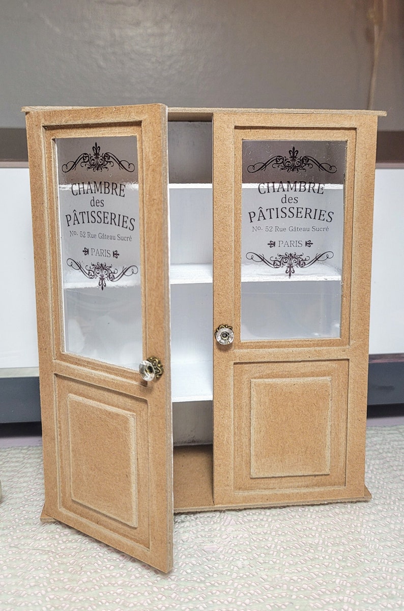 May include: A miniature wooden cabinet with two glass doors. The doors are open and feature a printed design that says "CHAMBRE des P&Acirc;TISSERIES No. 52 Rue G&acirc;teau Sucr&eacute; PARIS".