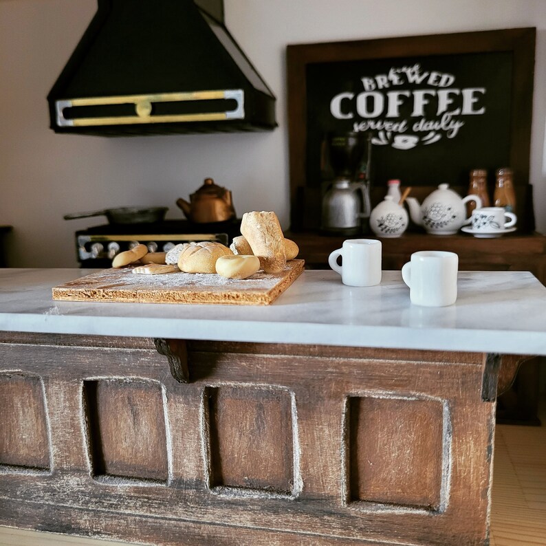 May include: A miniature kitchen scene with a wooden countertop and island. A wooden cutting board holds bread and a dusting of flour. Two white mugs sit on the counter. A chalkboard sign reads "BREWED COFFEE served daily".