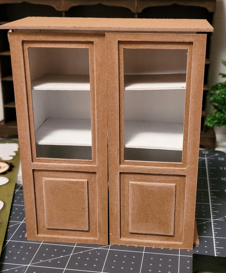 May include: A brown wooden cabinet with glass doors and two shelves. The cabinet has a simple design with a raised panel on each door.