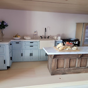 May include: Miniature kitchen scene with light blue cabinets, a white marble countertop, and a wooden island. Includes a black faucet, a stove, and decorative items like flowers, fruit, and bread. A charming dollhouse kitchen.