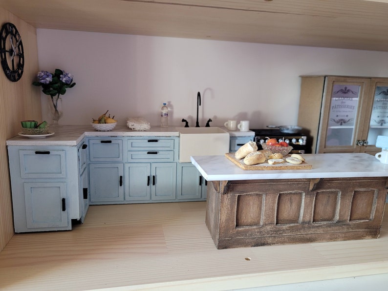 May include: Miniature kitchen scene with light blue cabinets, a white marble countertop, and a wooden island. Includes a black faucet, a stove, and decorative items like flowers, fruit, and bread. A charming dollhouse kitchen.