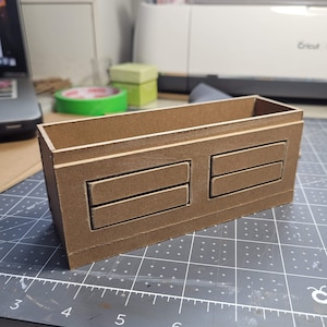 May include: A brown rectangular wooden box with two sets of two rectangular cutouts. The box is sitting on a gray cutting mat with a ruler. The box is likely for storage or organization.