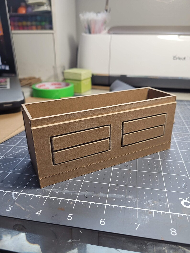 May include: A brown rectangular wooden box with two sets of two rectangular cutouts. The box is sitting on a gray cutting mat with a ruler. The box is likely for storage or organization.