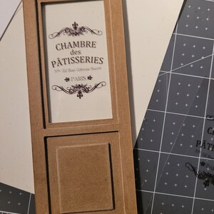 May include: A brown wooden frame with a glass pane displaying a sign that reads "CHAMBRE des PATISSERIES No. 32 Rue G&acirc;teau Sucr&eacute; PARIS". The frame has a raised panel below the glass pane.