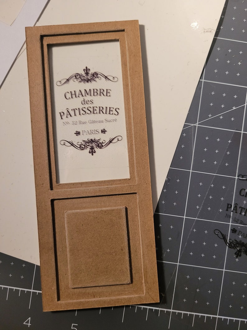 May include: A brown wooden frame with a glass pane displaying a sign that reads "CHAMBRE des PATISSERIES No. 32 Rue G&acirc;teau Sucr&eacute; PARIS". The frame has a raised panel below the glass pane.