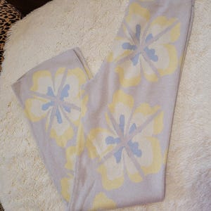 Puede incluir: Pantalones de punto lila claro con un estampado floral en amarillo y azul claro. Los pantalones est&aacute;n doblados y se muestran sobre una superficie blanca y esponjosa. El dise&ntilde;o floral es sim&eacute;trico y cubre la parte delantera.