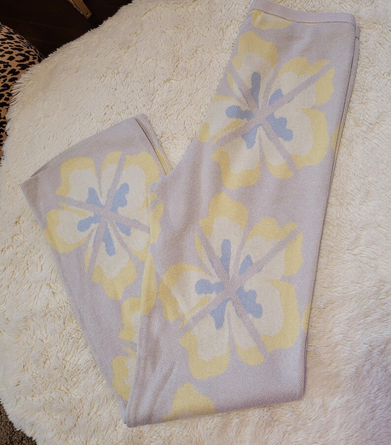 Puede incluir: Pantalones de punto lila claro con un estampado floral en amarillo y azul claro. Los pantalones est&aacute;n doblados y se muestran sobre una superficie blanca y esponjosa. El dise&ntilde;o floral es sim&eacute;trico y cubre la parte delantera.