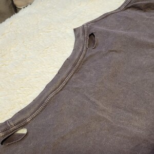 May include: Close-up of a faded purple sleeveless shirt with distressed details. The shirt has a worn, vintage look with visible holes near the shoulder and neckline. The fabric appears soft and well-worn, suggesting comfort and casual wear.