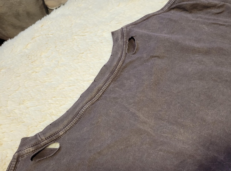 May include: Close-up of a faded purple sleeveless shirt with distressed details. The shirt has a worn, vintage look with visible holes near the shoulder and neckline. The fabric appears soft and well-worn, suggesting comfort and casual wear.