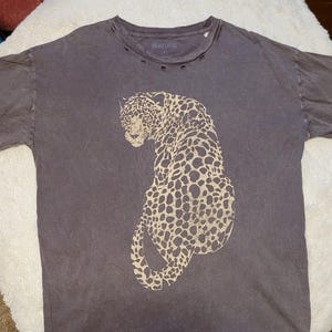 May include: A dark gray t-shirt featuring a large leopard design in tan and brown. The shirt has a distressed look with small holes near the neckline and short sleeves. The leopard is facing left, with its body curved.