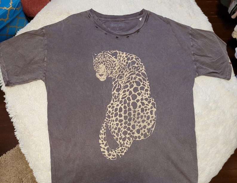 May include: A dark gray t-shirt featuring a large leopard design in tan and brown. The shirt has a distressed look with small holes near the neckline and short sleeves. The leopard is facing left, with its body curved.