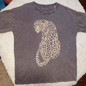May include: A dark gray t-shirt featuring a large, tan leopard print. The shirt has a crew neck with distressed detailing and short sleeves. The text "VINTAGE CULTURE" is printed on the neckline.