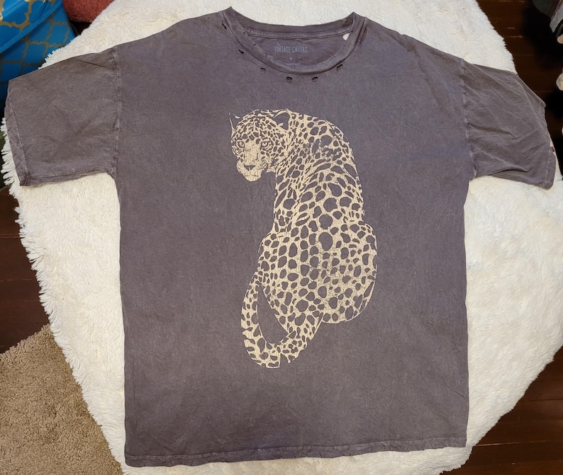 May include: A dark gray t-shirt featuring a large, tan leopard print. The shirt has a crew neck with distressed detailing and short sleeves. The text "VINTAGE CULTURE" is printed on the neckline.