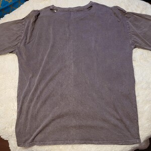 May include: A faded purple t-shirt with a crew neck and short sleeves. The shirt has a distressed look with small holes near the neckline. The fabric appears soft and worn, suggesting a vintage or pre-washed style.