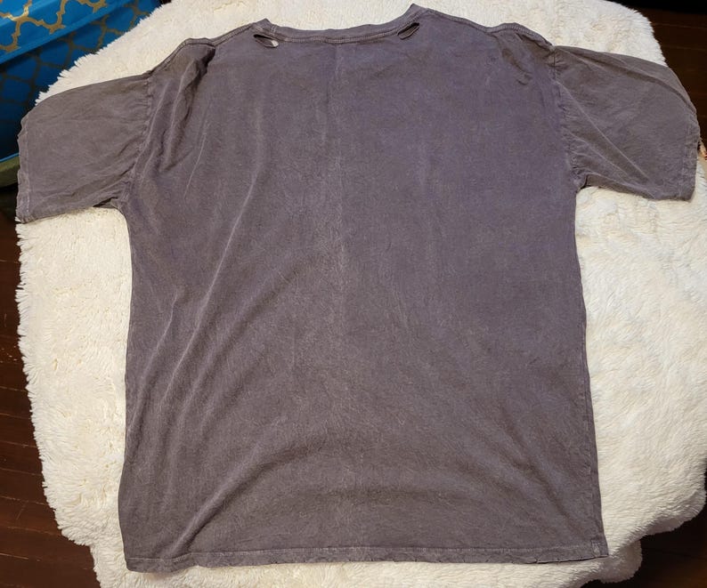 May include: A faded purple t-shirt with a crew neck and short sleeves. The shirt has a distressed look with small holes near the neckline. The fabric appears soft and worn, suggesting a vintage or pre-washed style.