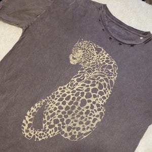 May include: A dark grey t-shirt featuring a large, tan leopard design. The shirt has a distressed look with small holes near the neckline and the text "VINTAGE CANVAS" printed on the collar.