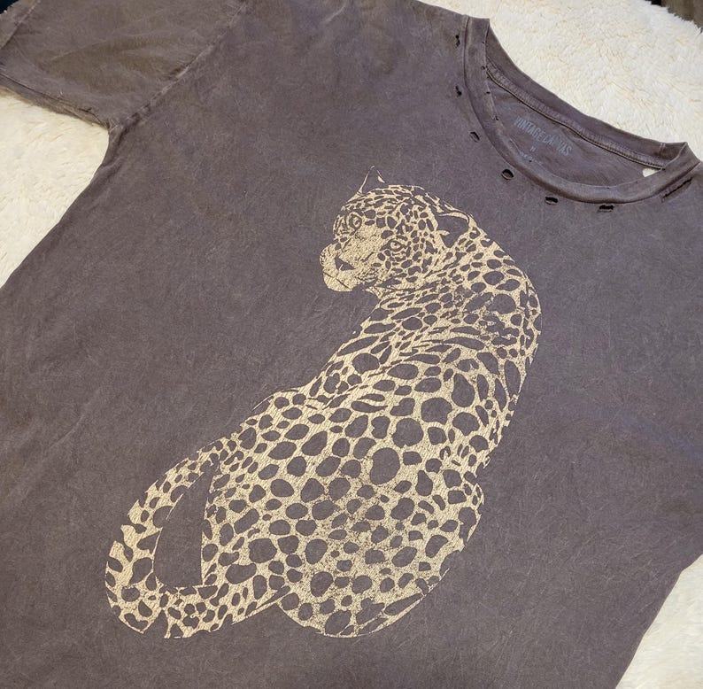 May include: A dark grey t-shirt featuring a large, tan leopard design. The shirt has a distressed look with small holes near the neckline and the text "VINTAGE CANVAS" printed on the collar.