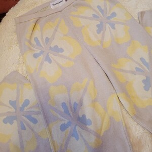 Puede incluir: Pantalones de punto beige claro con un gran estampado floral en amarillo y azul claro. Los pantalones est&aacute;n doblados y se muestran sobre una superficie suave y texturizada. La etiqueta dice "House of Sunny".