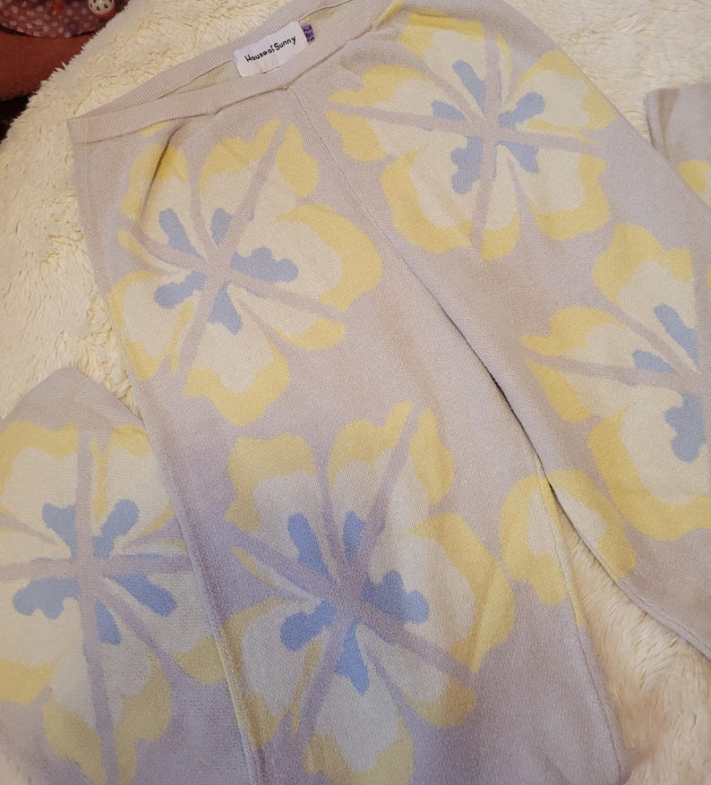Puede incluir: Pantalones de punto beige claro con un gran estampado floral en amarillo y azul claro. Los pantalones est&aacute;n doblados y se muestran sobre una superficie suave y texturizada. La etiqueta dice "House of Sunny".