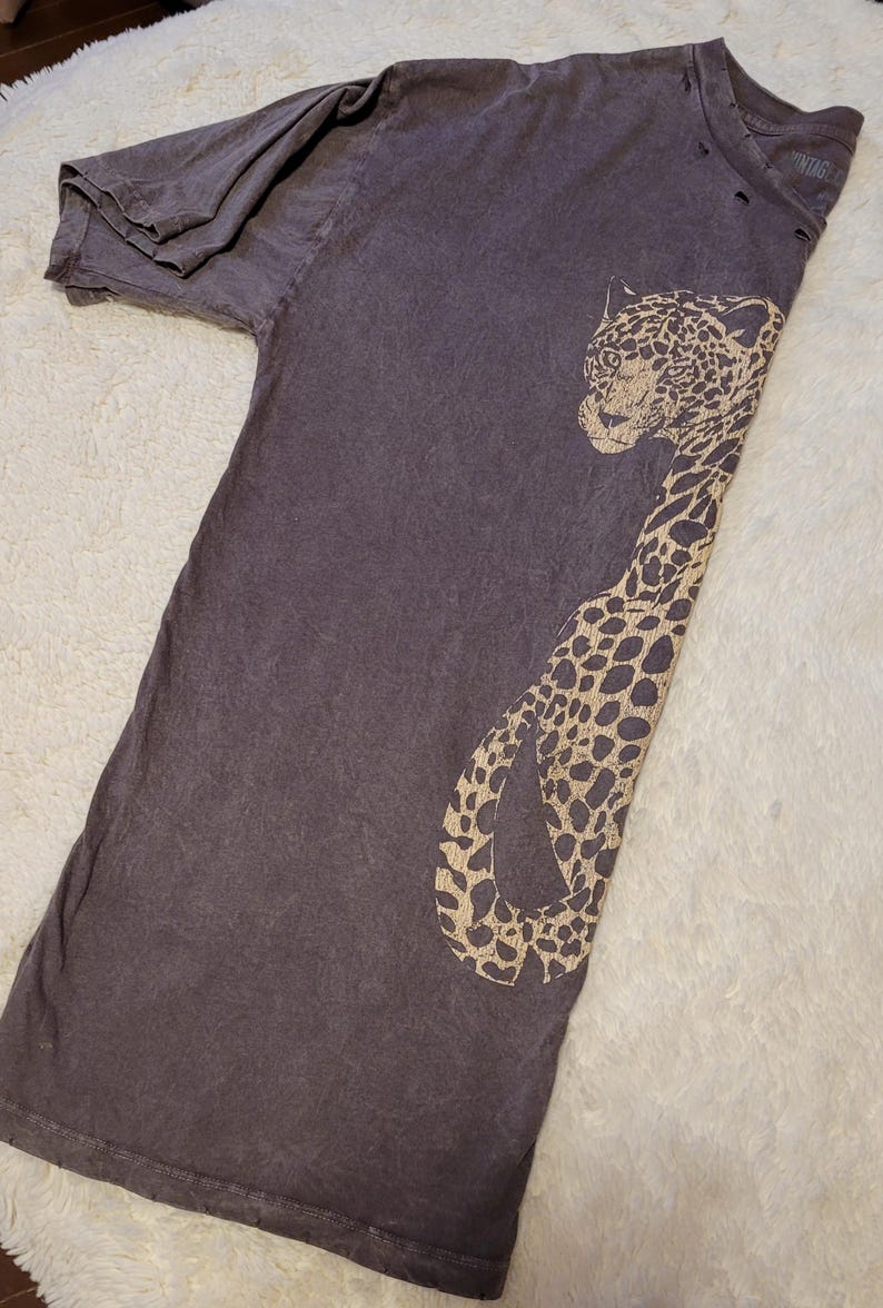 May include: A dark gray t-shirt featuring a tan leopard design. The distressed tee has a crew neck and short sleeves. The leopard graphic is on the front of the shirt.