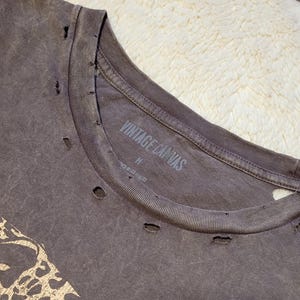 May include: A distressed, dark gray t-shirt with a crew neck and distressed holes. The shirt features the text "VINTAGE CANVAS" on the collar. The shirt has a medium size label.