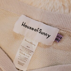 Puede incluir: Primer plano de un su&eacute;ter de punto beige con la etiqueta de la marca "House of Sunny" visible. La etiqueta incluye instrucciones de lavado e informaci&oacute;n de tallas. El su&eacute;ter est&aacute; hecho de 55% acr&iacute;lico y 45% algod&oacute;n.