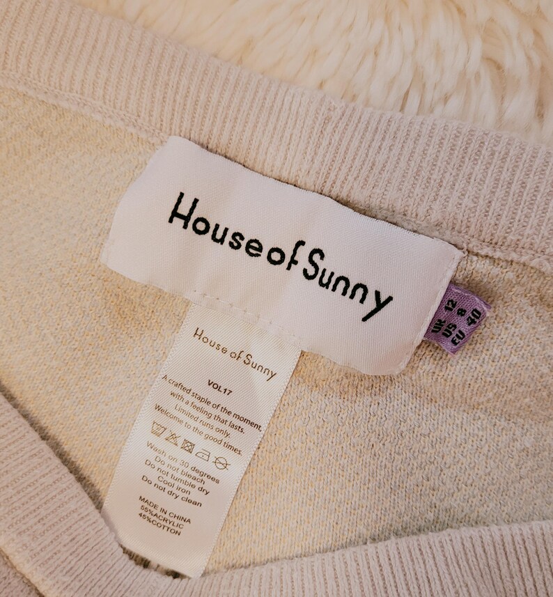 Puede incluir: Primer plano de un su&eacute;ter de punto beige con la etiqueta de la marca "House of Sunny" visible. La etiqueta incluye instrucciones de lavado e informaci&oacute;n de tallas. El su&eacute;ter est&aacute; hecho de 55% acr&iacute;lico y 45% algod&oacute;n.