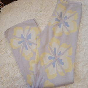 Puede incluir: Pantalones de punto lila claro con grandes estampados florales en amarillo y azul claro. Los pantalones est&aacute;n doblados y descansan sobre una alfombra blanca y esponjosa. El dise&ntilde;o floral est&aacute; distribuido uniformemente.