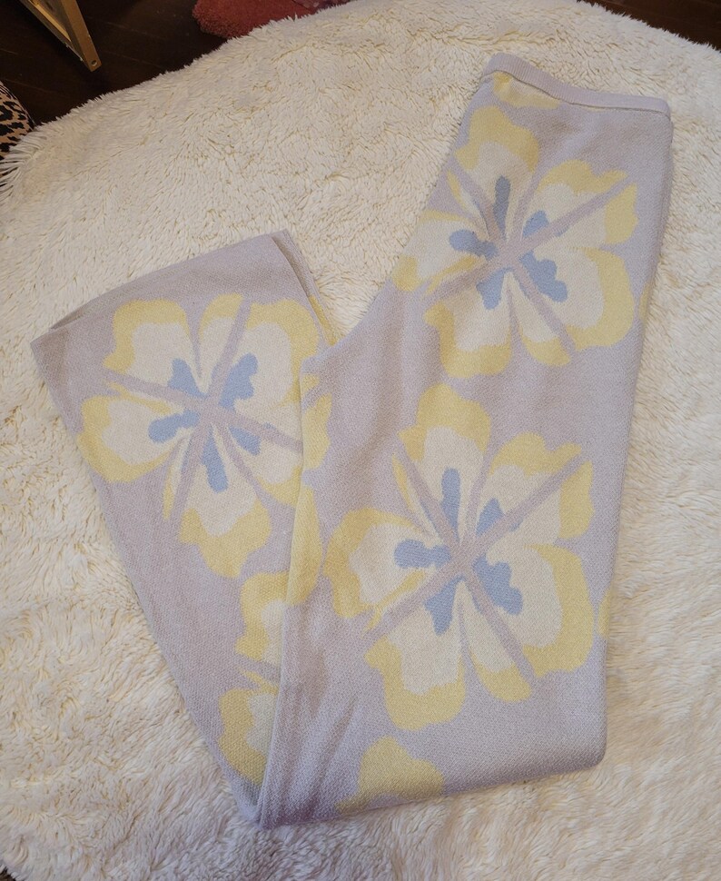 Puede incluir: Pantalones de punto lila claro con grandes estampados florales en amarillo y azul claro. Los pantalones est&aacute;n doblados y descansan sobre una alfombra blanca y esponjosa. El dise&ntilde;o floral est&aacute; distribuido uniformemente.
