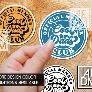 May include: Two round stickers with the text "Official Member Cool Dads Club" in a retro font. The stickers are in blue and yellow with black text. The stickers are on a wooden surface.