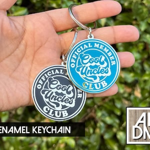 May include: Two enamel keychains with the text "Cool Uncles Club" in a circular design. One keychain is black and white, the other is blue and white. The keychains are held up against a green background.