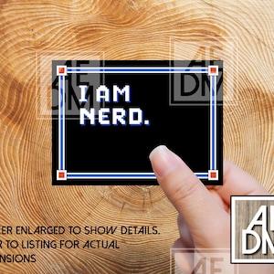 May include: A black sticker with the text "I AM NERD." in white pixelated font, framed by a blue and red border. The sticker is held against a wooden background.