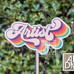 May include: A die-cut sticker featuring the word Artist in a bold, cursive font. The lettering is layered over a series of concentric, wavy rainbow-colored stripes in shades of purple, red, orange, yellow, and teal, creating a retro-inspired design.