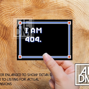 May include: A black sticker with the text "I AM 404." in white pixelated font, framed by a blue border with red corner accents. The sticker is held by a hand and placed on a wooden surface.
