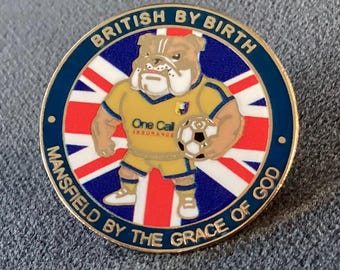 May include: A round metal pin featuring a bulldog wearing a yellow football kit with the text One Call Insurance. The bulldog holds a football against a Union Jack background. The border text reads BRITISH BY BIRTH MANSFIELD BY THE GRACE OF GOD.