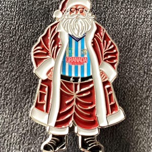 May include: A metal enamel pin featuring Santa Claus wearing a red and white coat over a light blue and white striped shirt with the text GRANADA BINGO printed in red on a white rectangle, paired with red trousers and black boots.