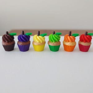 May include: A row of six miniature cupcake figurines in brown, purple, yellow, green, orange, and red. Each cupcake features a swirled frosting top, a small dark brown stem, and a single green leaf, all resting in matching colored fluted paper liners.