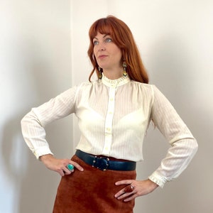 Può includere: Una donna indossa una camicia bianca con colletto e colletto arricciato, una gonna di camoscio marrone, una cintura nera e stivali marroni. È in piedi in una stanza con pareti bianche.