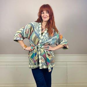 May include: A quilted jacket featuring a patchwork design with various muted colours including sage green, tan, and blue. The jacket has three-quarter length sleeves, a wrap-style front with a matching fabric tie belt, and a V-neckline.