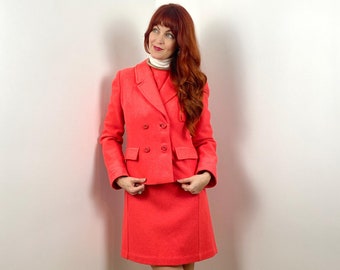 May include: A woman wearing a bright pink double-breasted suit with a white turtleneck and red loafers. She is standing in a corner with a white wall behind her.