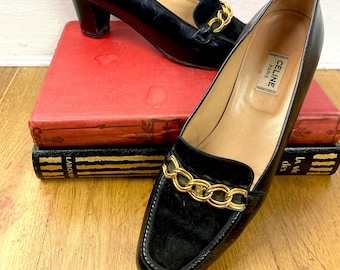 Könnte beinhalten: Schwarze Leder-Slipper mit goldenem Kettendetail und einem dicken Absatz. Die Schuhe stehen auf einem Stapel Bücher. Das Etikett auf dem Schuh lautet "CELINE PARIS."