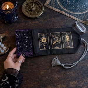 May include: A black fabric tarot card roll featuring a purple starry velvet flap and three gold-printed cards labelled The Sun, The Chariot, and The Moon. The roll includes a grey braided cord with a matching tassel for secure closure.