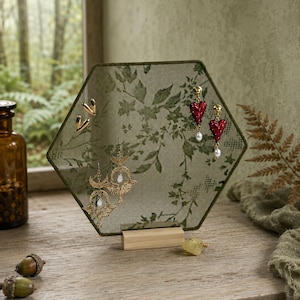 May include: A hexagonal jewelry display stand featuring a green mesh screen with a dark green leaf pattern, supported by a light wood base. The stand holds gold-toned heart studs, red heart-shaped drop earrings with pearls, and gold crescent moon earrings.