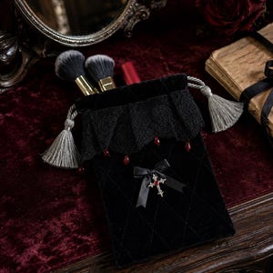 Puede incluir: Una bolsa de terciopelo negro con cordón, patrón de diamantes acolchado, ribete de encaje negro con pequeños detalles de gotas rojas y un lazo de cinta negra con un dije de cruz plateada. La bolsa incluye dos borlas de color gris.