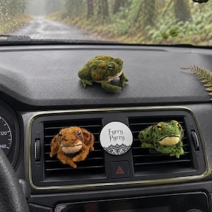 May include: Three needle-felted frog figures in green and brown tones. One frog sits on the dashboard, while two others are attached to the car air vent. A circular white disc with black lace trim and the text Fyrry Pyrry is between the vent frogs.