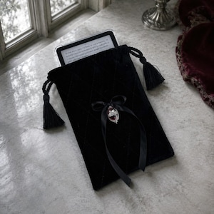 May include: A black quilted velvet drawstring pouch featuring a black ribbon bow and a silver-toned pendant with a dark red stone. The pouch contains an e-reader with visible text, and the closure is secured with two black tassels on cords.